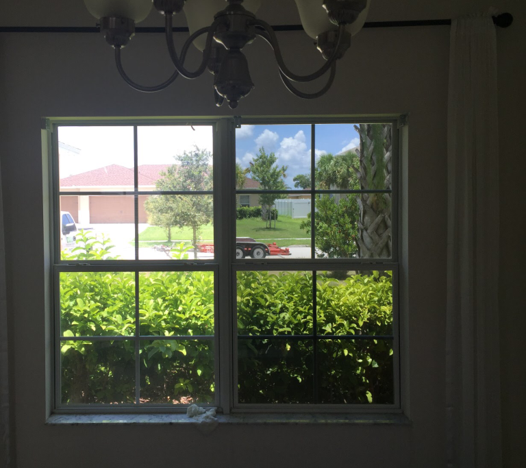 View through a window with symmetrical grids, showing lush greenery, a trailer parked on the street, and a house with red roof under a sunny sky.