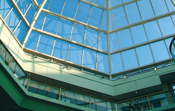 Mall atrium with a geometric glass ceiling, creating an open, airy space. Bright blue sky visible, conveying a modern and spacious atmosphere.