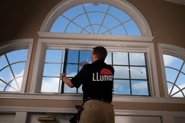 A person in a black "LLumar" shirt installs window film on a large, semi-circular window. Bright blue sky visible outside, creating an airy, clean atmosphere.