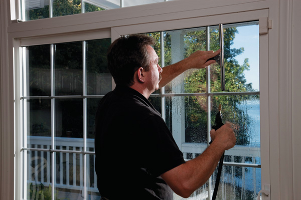A person cleaning a large glass door with a squeegee. Sunlight illuminates the space, highlighting a scenic view of trees and water outside. Calm and focused setting.