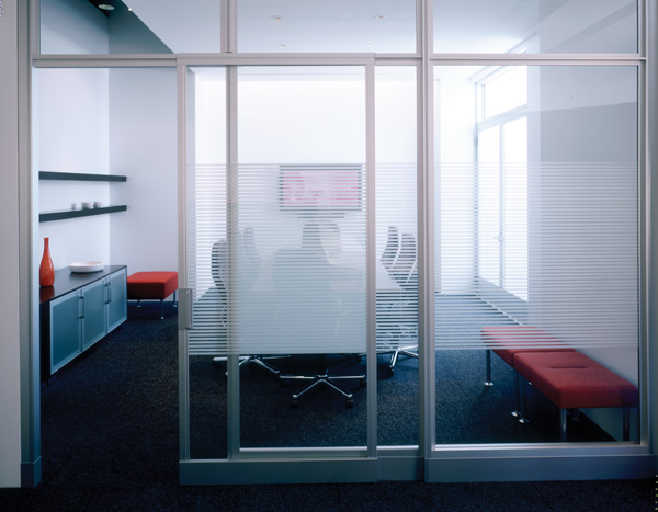 Modern conference room with frosted glass walls and a table surrounded by chairs. Shelves and red accents create a sleek, professional atmosphere.