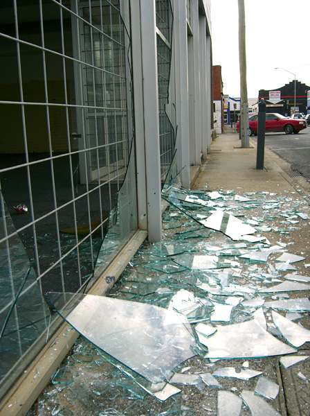 Shattered glass litters a sidewalk beside a building with metal grates, conveying a sense of disruption. A red car is visible on the street.
