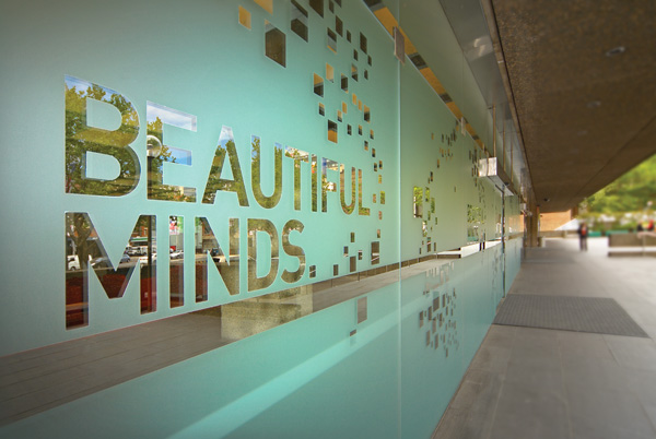 Glass wall with reflective silver letters spelling "BEAUTIFUL MINDS." Surrounding small square patterns. Modern, contemplative exterior scene.